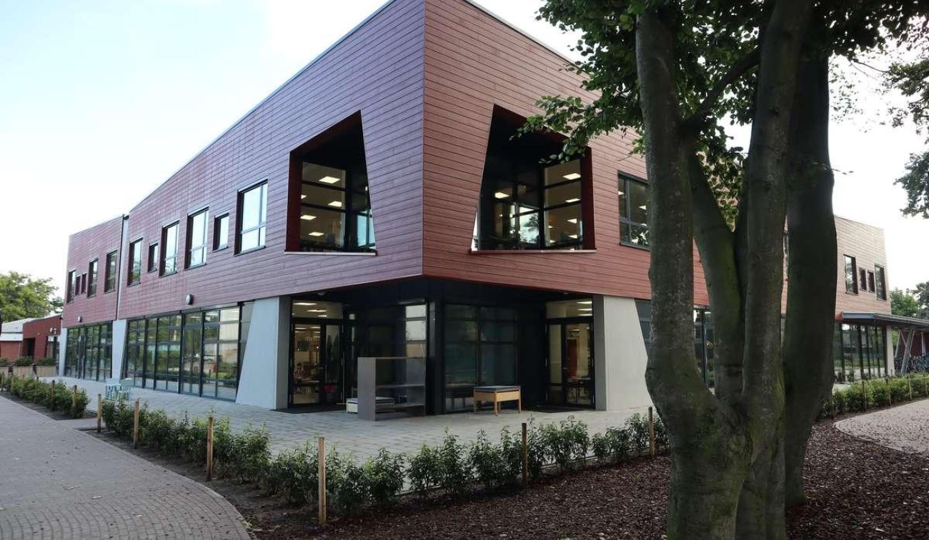 Hoekaanzicht van de nieuwbouw van basisschool Den Doelhof in Meijel met rood gevelhout, grote schuine raamkaders en glazen plint; ernaast een slingerend pad met jonge beplanting en bomen.