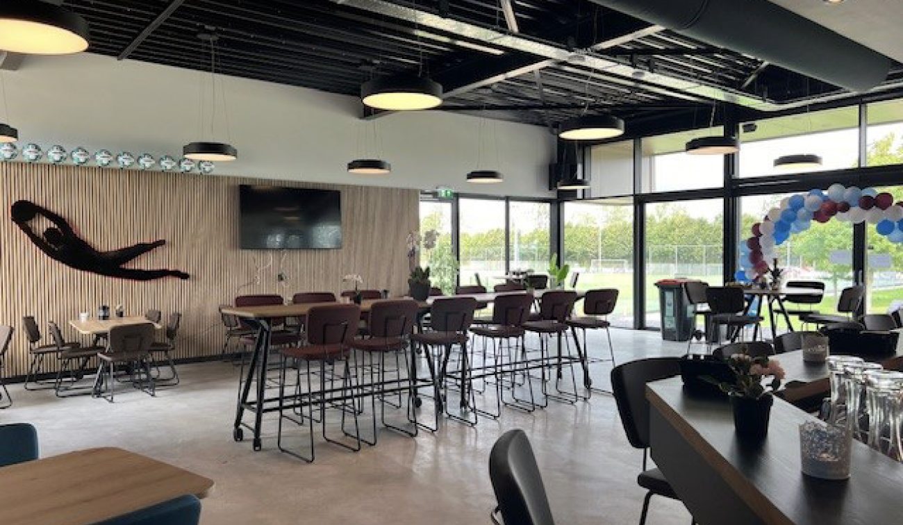 Clubkantine FC Landgraaf: hoge tafels, hanglampen, lamellenwand met tv en glas naar de velden