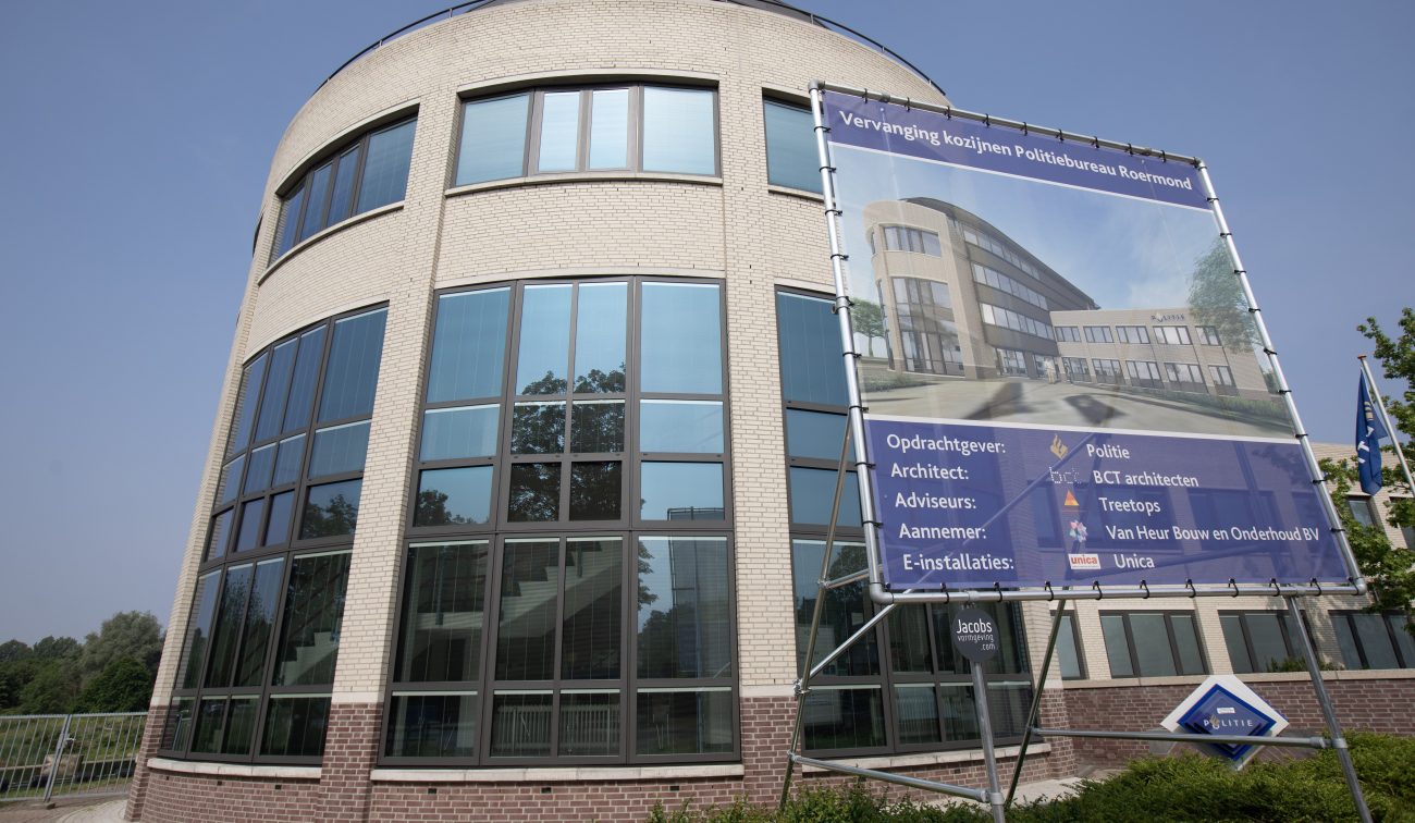 Close-up van de ronde glazen gevel van het politiebureau in Roermond met ervoor een groot projectbord over de vervanging van kozijnen (met opdrachtgever, architect en aannemer vermeld).