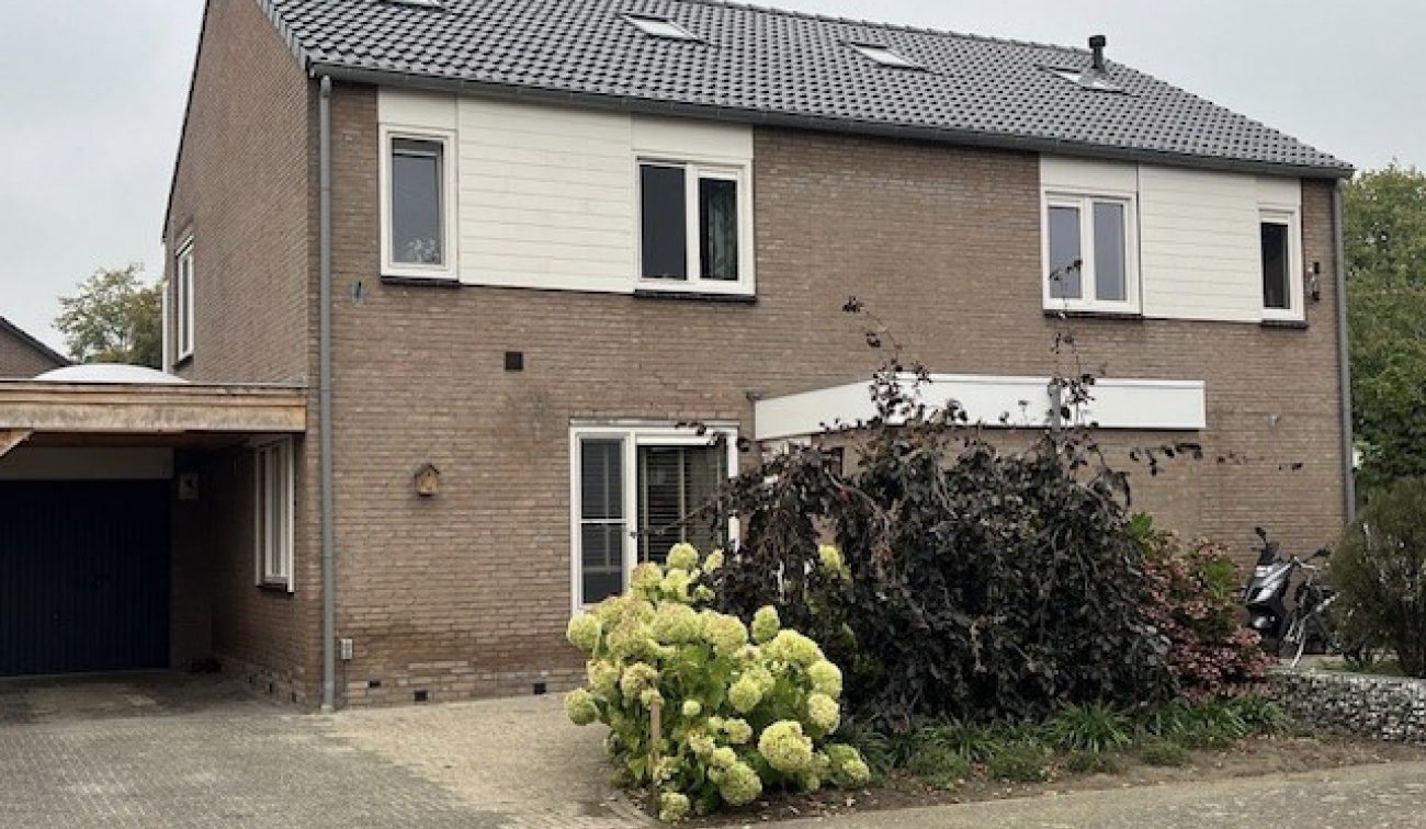 Twee-onder-een-kapwoning in Stramproy (project Groot onderhoud 28 woningen): bakstenen gevel, grijs pannendak met vier dakramen en witte geveldelen op de verdieping; oprit/carport links en voortuin met hortensia op een bewolkte dag.
