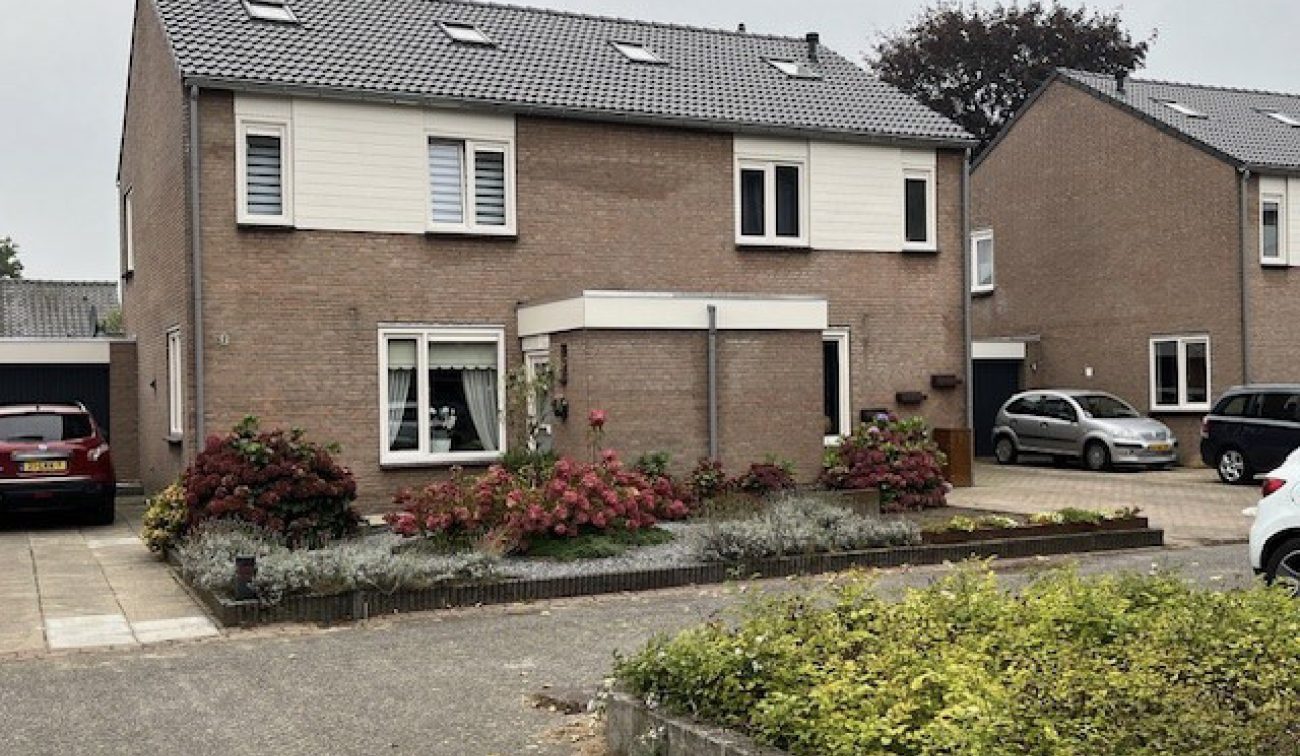 Twee-onder-een-kapwoningen in Stramproy (groot onderhoud) met bakstenen gevels, grijze pannendaken en witte geveldelen op de verdieping; entree-uitbouw in het midden, verzorgde voortuin met struiken en opritten met geparkeerde auto’s onder grijze lucht.