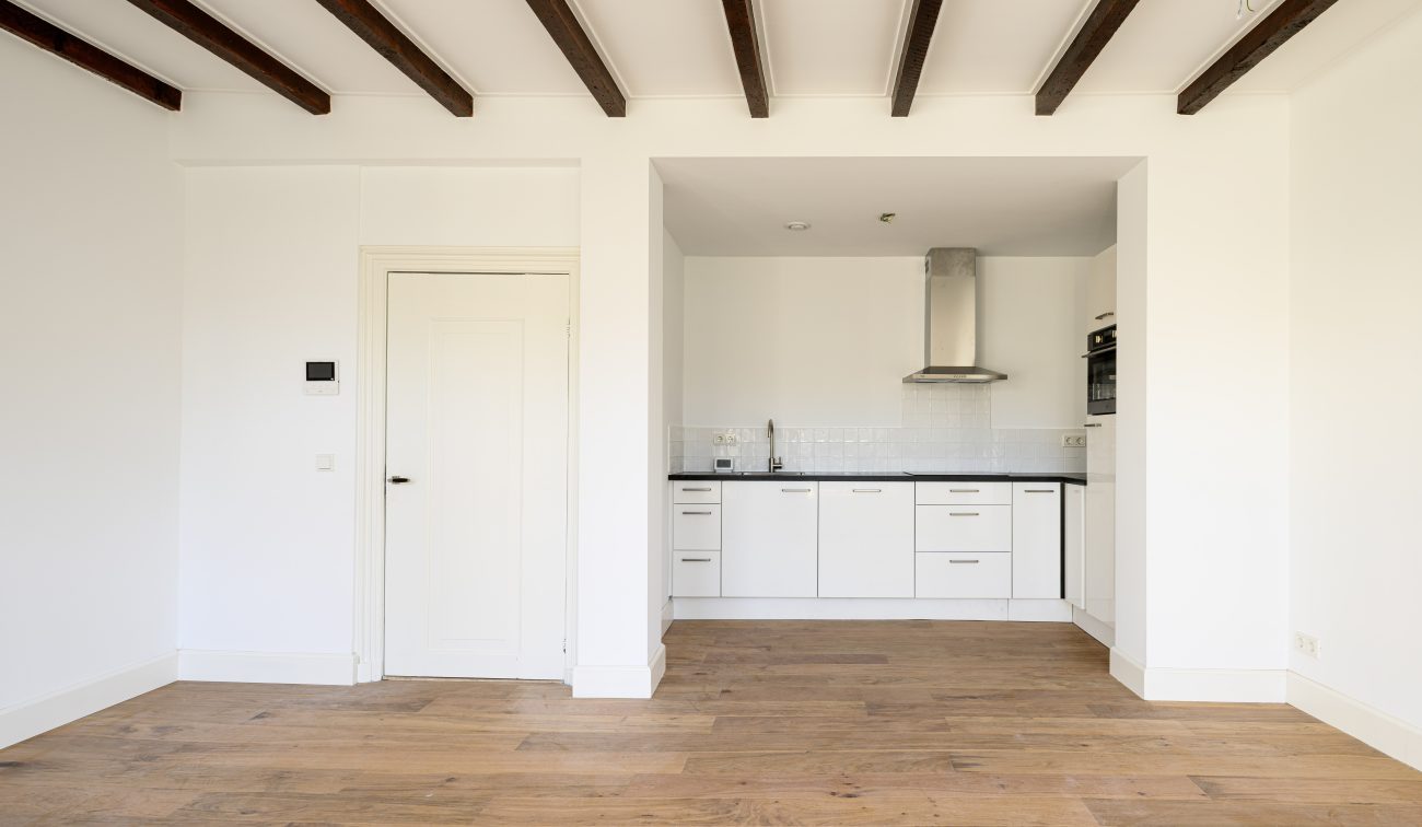 Transformatie Lijnmarkt & Visschersplein Utrecht – woonkamer met open keuken in nis, witte kasten en rvs-afzuigkap, zichtbare balken en eikenhouten vloer.