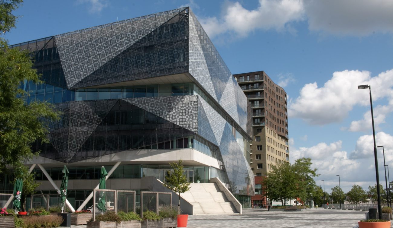 Hoekig glazen volume boven transparante plint; plein met plantenbakken en toren op achtergrond.