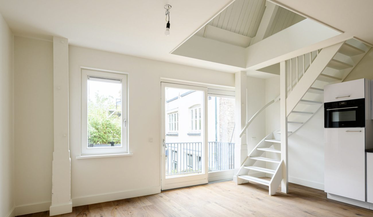 Renovatieappartement: open trap, wit balkenplafond, schuif-/tuindeur met frans balkon en kolomkast met oven.