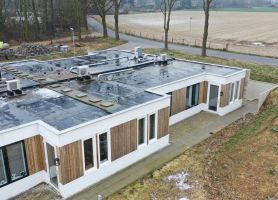 Hoekaanzicht nieuwbouw zorgwoning Amstenrade met witte en houten gevel, plat dak vol installaties, betegeld pad langs de gevel en landelijke omgeving met weg en velden.