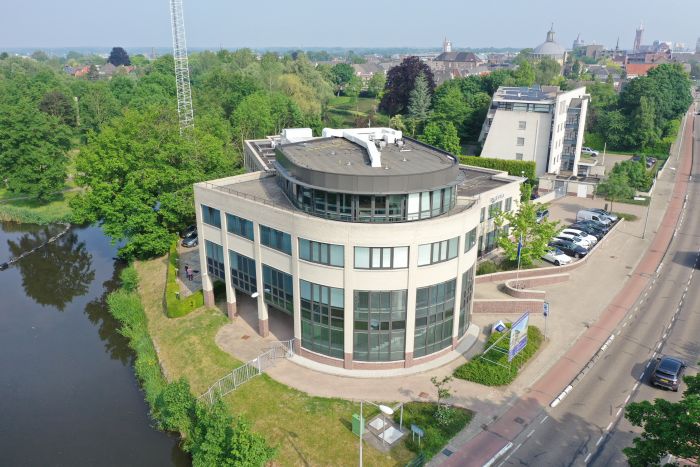 Luchtfoto van het gerenoveerde politiebureau in Roermond met ronde glazen hoekgevel, donkergrijze dakopbouw en plat dak, gelegen aan het water met parkeervakken en toegangsweg rechts.