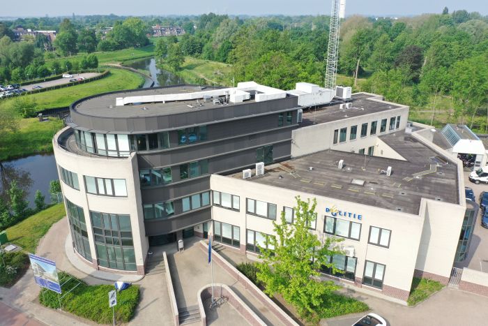 Luchtfoto van het politiebureau in Roermond met ronde glazen hoek en donkere bovenbouw, zicht op platte daken met installaties en het ‘POLITIE’-logo aan de gevel, omringd door groen en water.