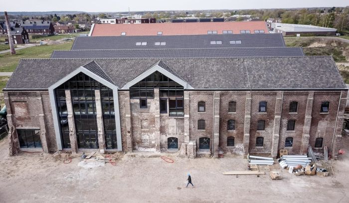 Voorgevel van een gerestaureerde bakstenen fabriek met zadeldak; twee grote glazen puien in de geveldelen links, verweerde metselwerkvakken met boogramen rechts, en bouwmaterialen/werkterrein op de voorgrond.