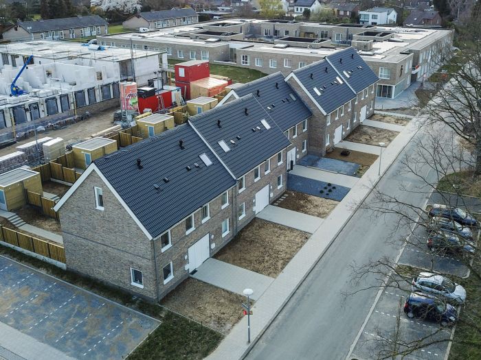 Nieuwbouw rij eengezinswoningen in Hulsberg met bakstenen gevels en donkere pannendaken; nieuwe stoepen en opritten, houten tuinen/bergingen en bouwplaats links.