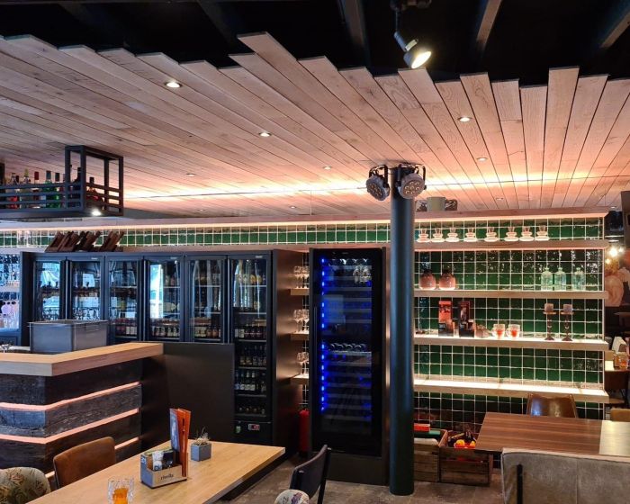 Interieur eetcafé De Aanwinst: barwand met koelvitrines en open schappen met groen tegelwerk, houten plafond en staande spot.