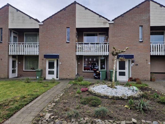 Portiekwoningen in Stramproy (Groot onderhoud 28 woningen) met bakstenen gevel, witte balkonhekken en blauwe luifels; twee entrees met stoep en groencontainers, voortuin met grindcirkel en gesnoeide boom, scooter achter raam zichtbaar.