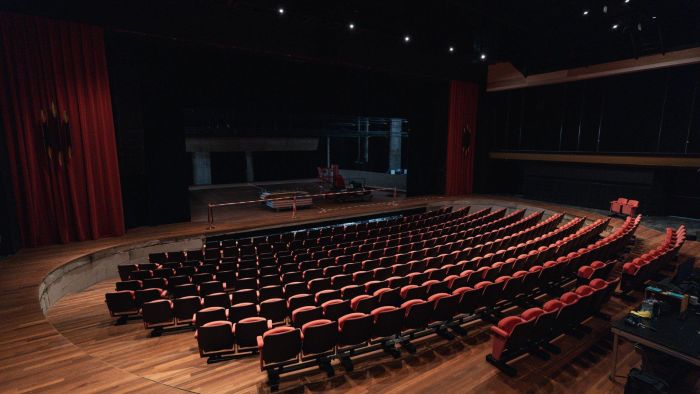 Theaterzaal in verbouwing met gebogen rijen rode stoelen, houten vloer en podium met bouwmaterialen; rode gordijnen en gedimde verlichting in Theaterhotel De Oranjerie, Roermond.