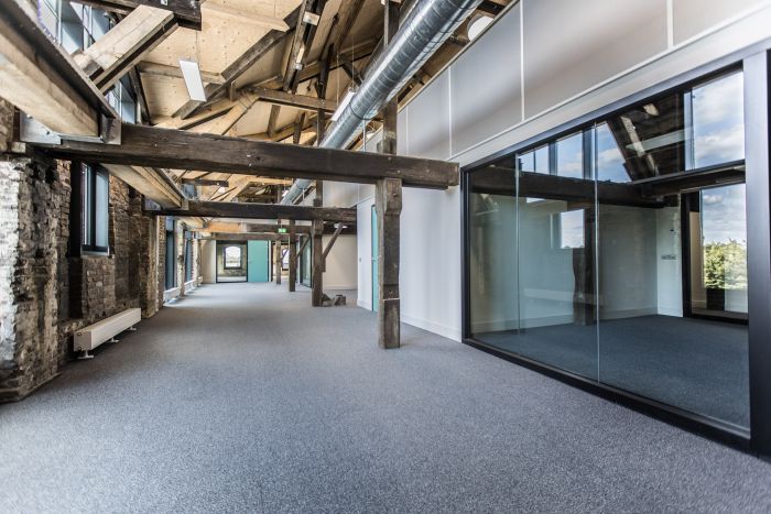 Gerestaureerd fabriekinterieur met zichtbare houten balken en metselwerk, grijs tapijt, glazen scheidingswand rechts en zichtbare luchtkanalen onder het schuine dak, met daglicht tot aan het eind van de gang.