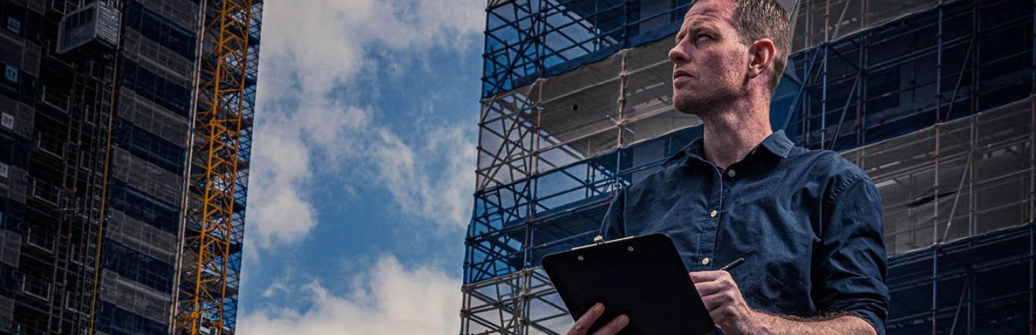 Man met een clipboard bekijkt een bouwplaats met steigers en hoge gebouwen op de achtergrond.