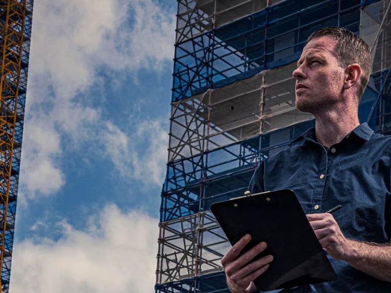 Man met een clipboard bekijkt een bouwplaats met steigers en hoge gebouwen op de achtergrond.